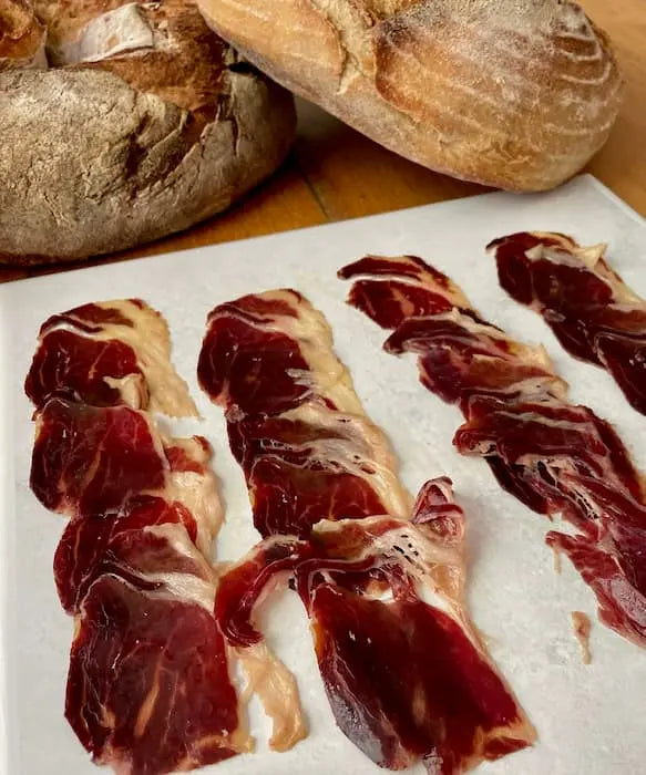 Finas rebanadas de Paleta ibérica 100% de bellota de 5-5,5 kg se disponen sobre una superficie blanca, con dos panes rústicos de fondo sobre una mesa de madera.