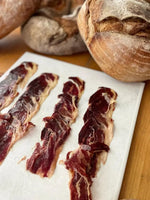 Tres rebanadas de Paleta ibérica 100% de bellota de 5-5,5 kg están dispuestas sobre una pizarra blanca, con pan rústico redondo de fondo sobre una superficie de madera.