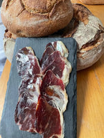 Sobre una superficie de madera, finas lonchas de Paleta ibérica 100% de bellota de 5-5,5 kg se disponen en un plato de pizarra negra frente a dos rústicas hogazas de pan crujiente.