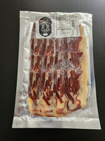 Un envase al vacío de Paleta ibérica 100% de bellota de la Sierra de Aracena-Jabugo, en lonchas finas, con etiqueta visible y carne roja y blanca entreverada perfectamente dispuesta dentro de un envase transparente sobre una superficie oscura.